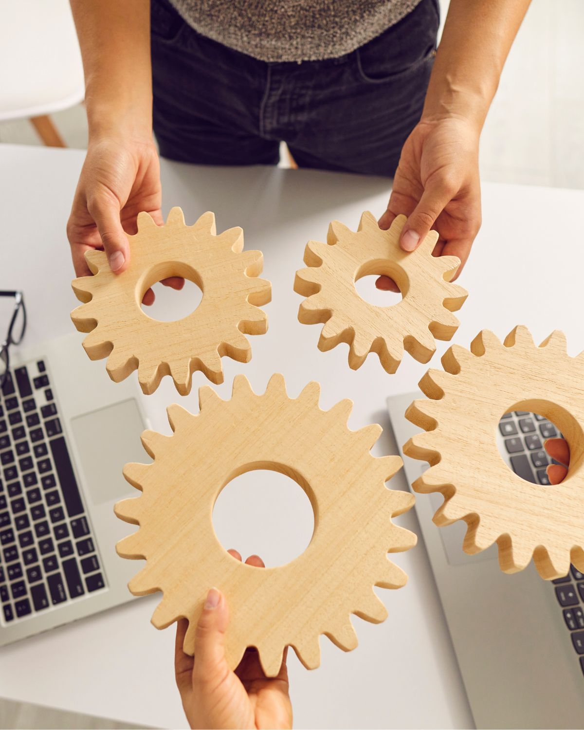 Top down view of a table with a computer and 2 people holding wooden gears tin an abstract way of showing problem solving.